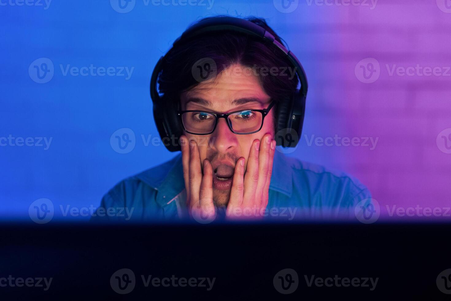This image depicts a young man sitting in front of a computer screen, wearing headphones. He has a shocked expression on his face, with his hands covering his mouth. photo