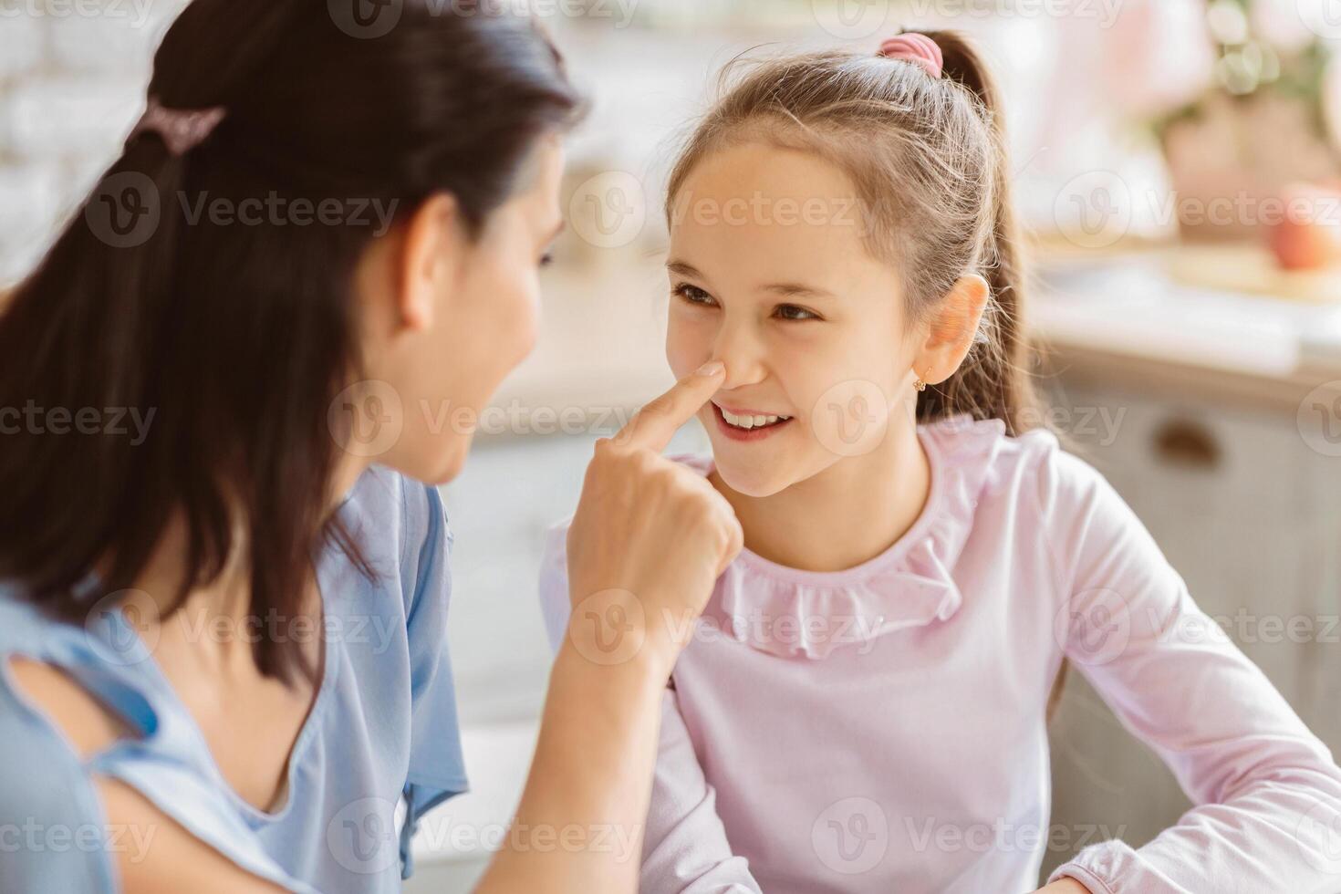 A mother and her young daughter are playing a game where they touch each others noses. The girl is smiling and looking at her mother with happiness. They appear to be in their kitchen photo