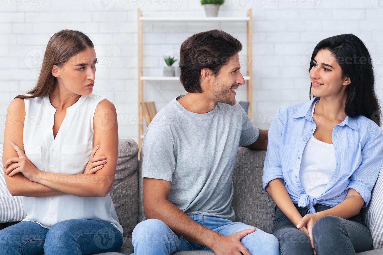 Jealousy. Angry Girlfriend Looking At Boyfriend Flirting With Another Girl Sitting On Sofa Indoor. photo