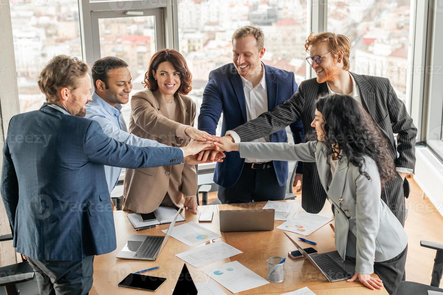A diverse group of business professionals stand around a conference table, their hands stacked together in a gesture of teamwork and unity. The office is modern and bright photo