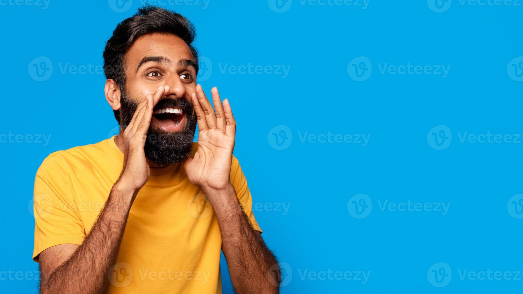 Indian young man with a beard is standing in front of a blue background. He is cupping his hand around his mouth and shouting, copy space. He has a yellow t-shirt on and appears happy and excited. photo