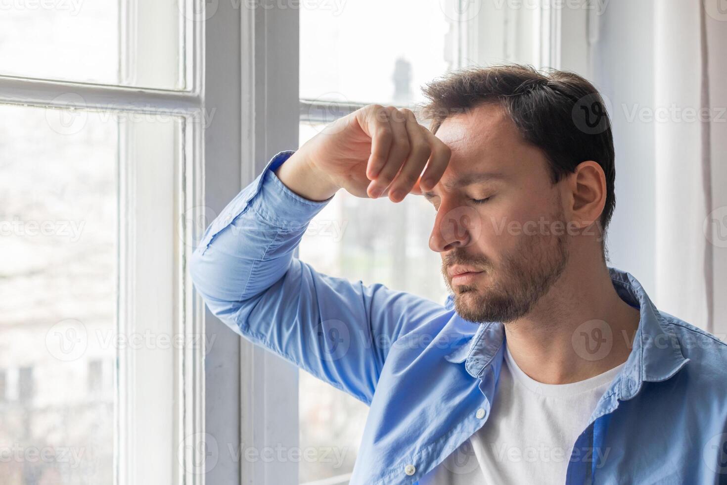 A man standing in front of a window, holding his head with his hands. His facial expression shows signs of distress or deep thought as he looks outside. photo