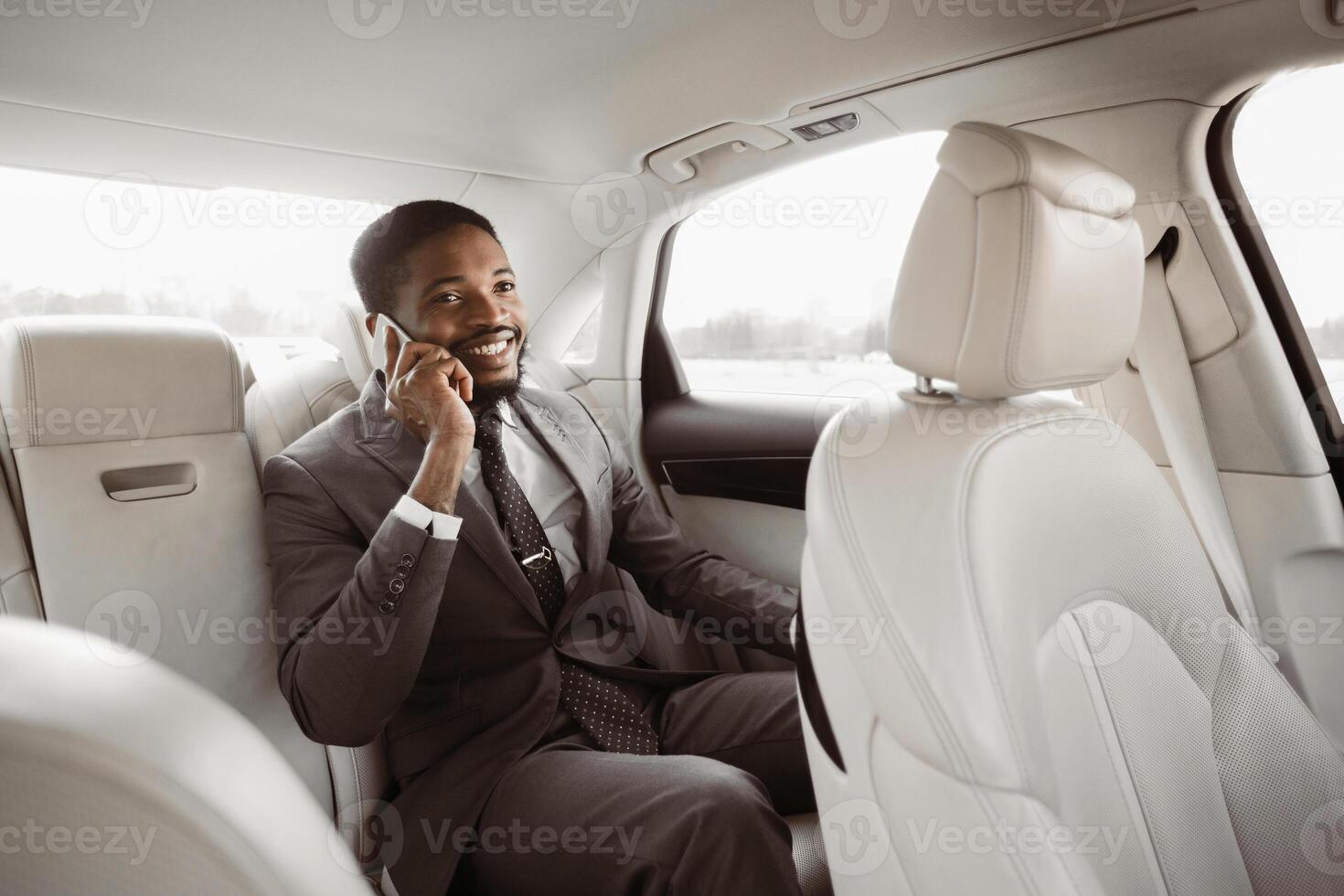 Black businessman in a suit is sitting in the back seat of a car while talking on his phone. He has a serious expression on his face and is looking to the side photo