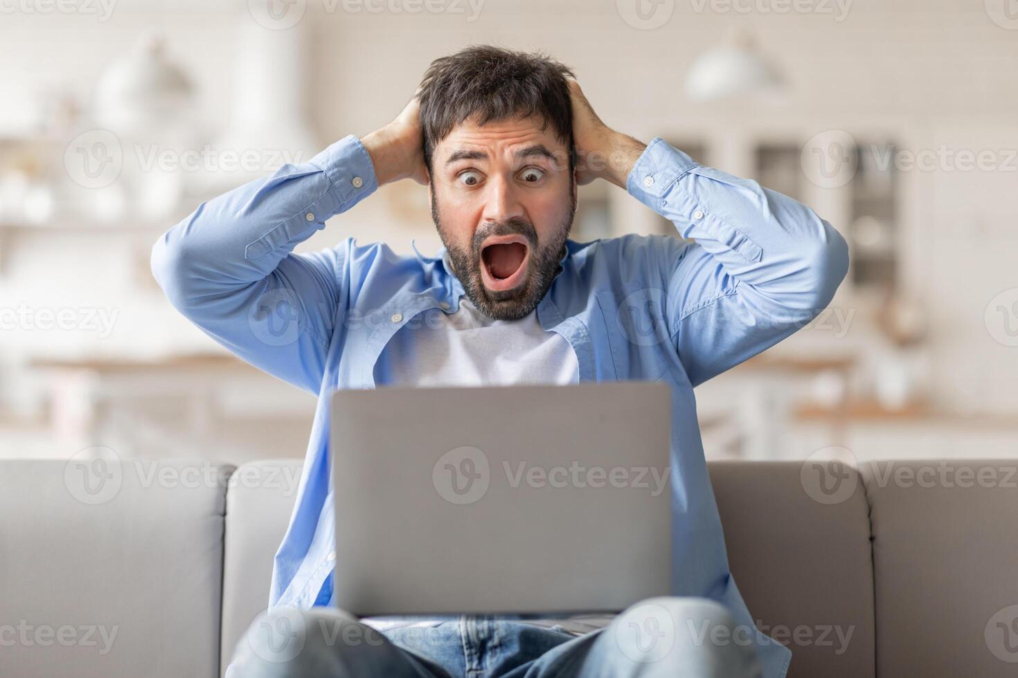 Man sitting on a couch, staring in shock at his laptop screen. His hands are on his head, and his mouth is open in surprise. He appears to be reacting to something he has just seen on the screen photo