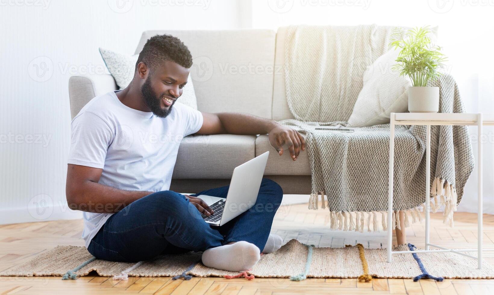 A young black man sits on the floor of his living room, using a laptop computer. He is sitting on a rug in front of a couch, with a plant on a side table photo