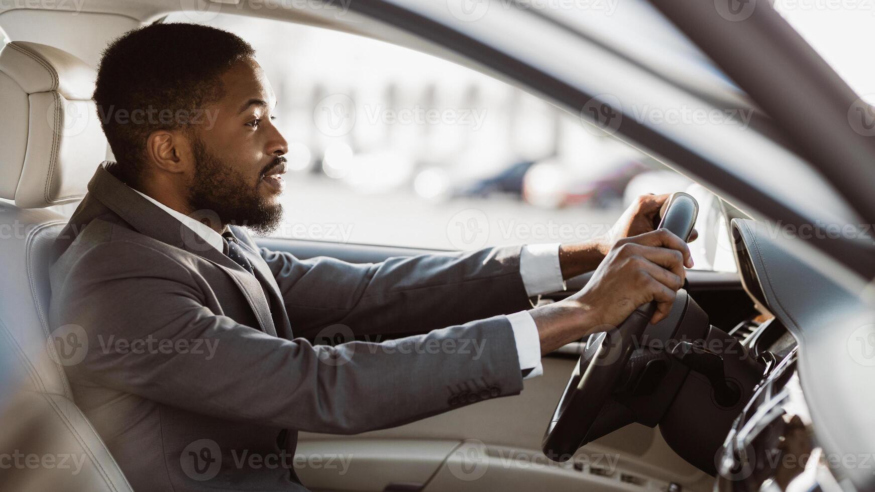 A professional black man wearing a suit and tie is driving a car. He is looking out the window to the right of the frame. The sun is shining brightly, making the inside of the car look bright. photo