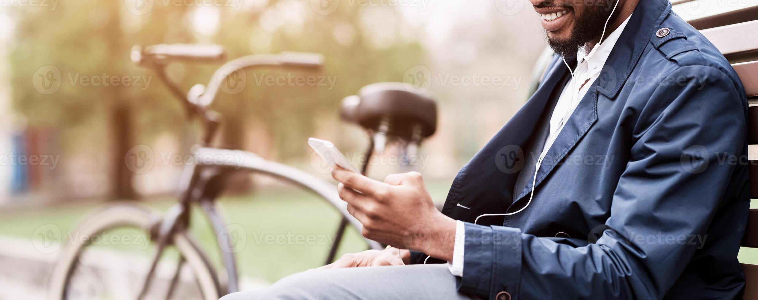 A businessman is seated on a park bench, using his smartphone, with a bicycle nearby. The environment is serene and professional, showcasing a balance between work and nature, cropped photo