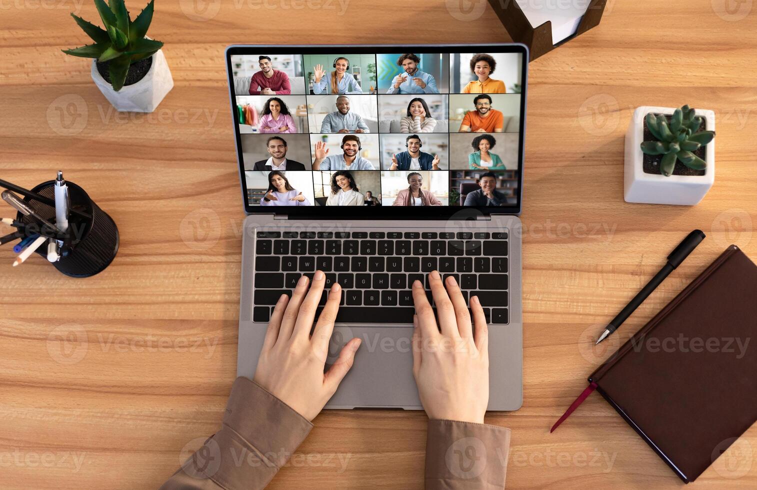 A person is participating in a conference using a laptop computer. The screen shows multiple people in a grid format. The persons hands are on the keyboard of the laptop, top view photo