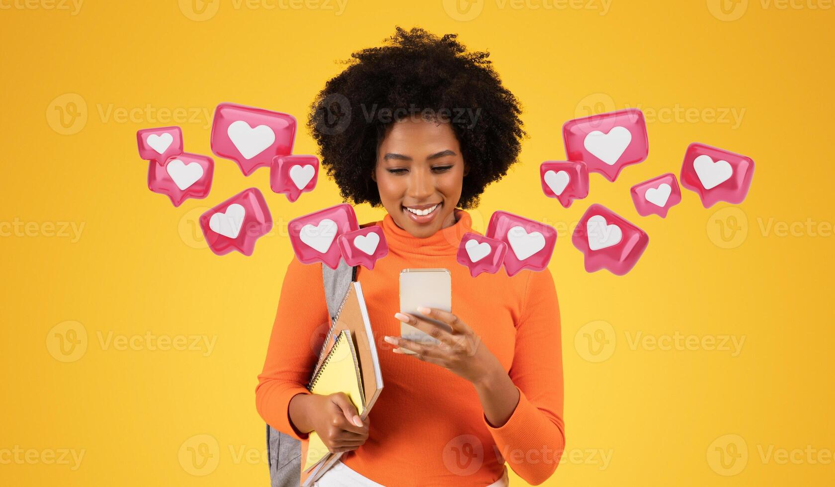 Engaged young woman with curly hair, wearing an orange sweater, looking at her smartphone with a smile, texting lover, holding notebooks, with a grey backpack, against a yellow background photo