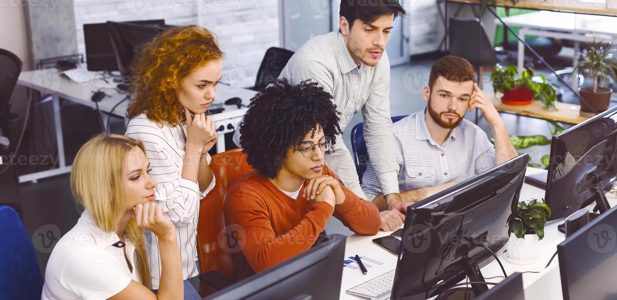 A group of focused multiethnic colleagues are seated at desks using desktop computers in a well-lit, contemporary office. They appear to be engrossed in work, displaying concentration and teamwork photo