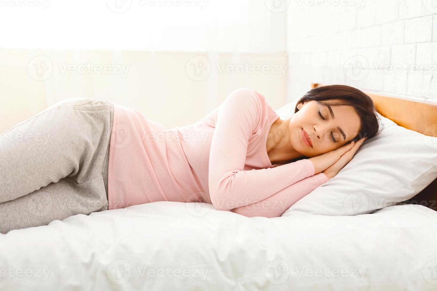 A young woman is captured in a serene moment of rest, lying comfortably on a plush white bed. She is peacefully napping, with her hands gently resting beside her head, her expression relaxed photo