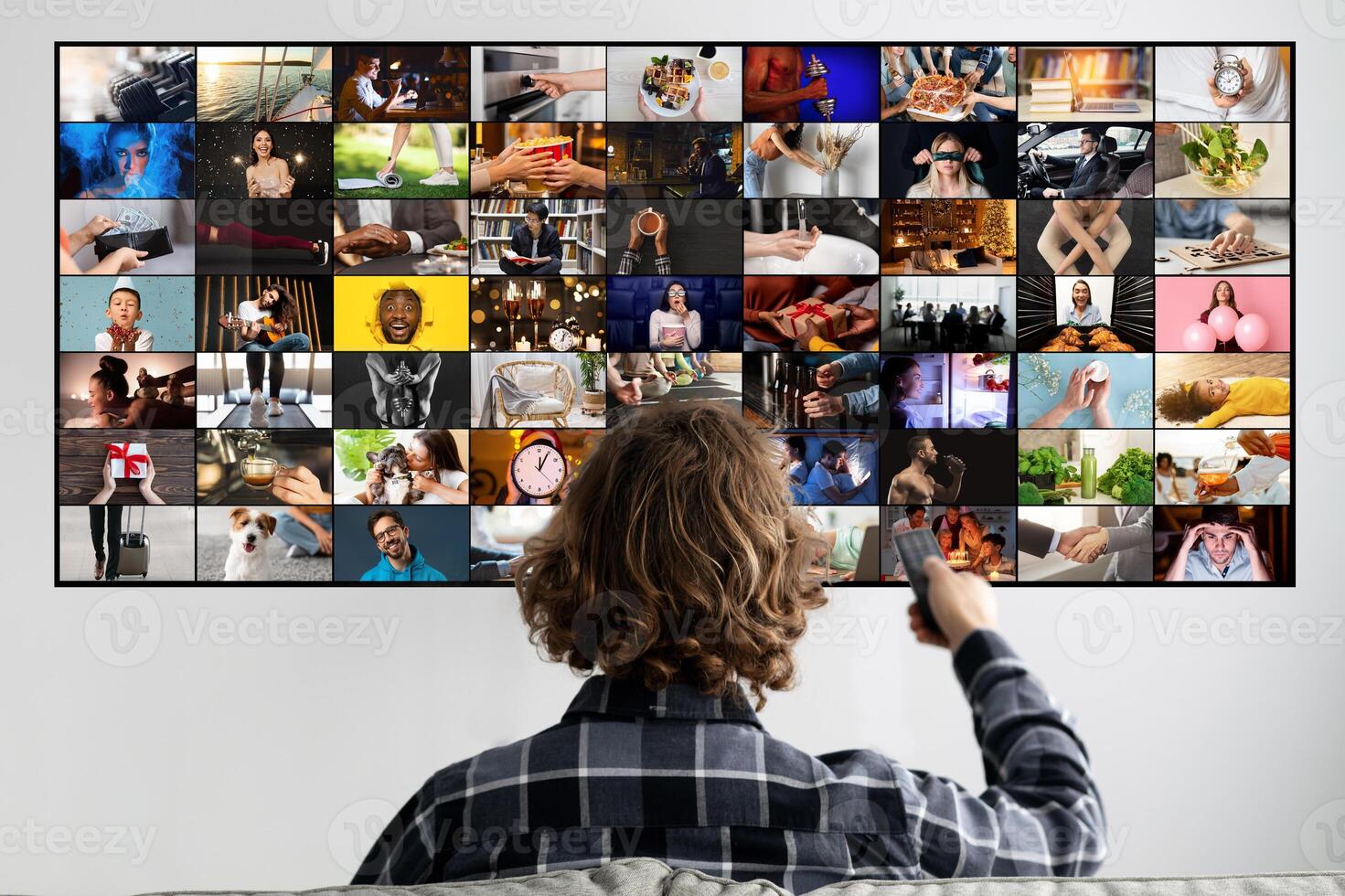 This image shows a man sitting in front of a large television screen. The television is displaying a grid of multiple small videos. The man is holding a remote control in his right hand photo