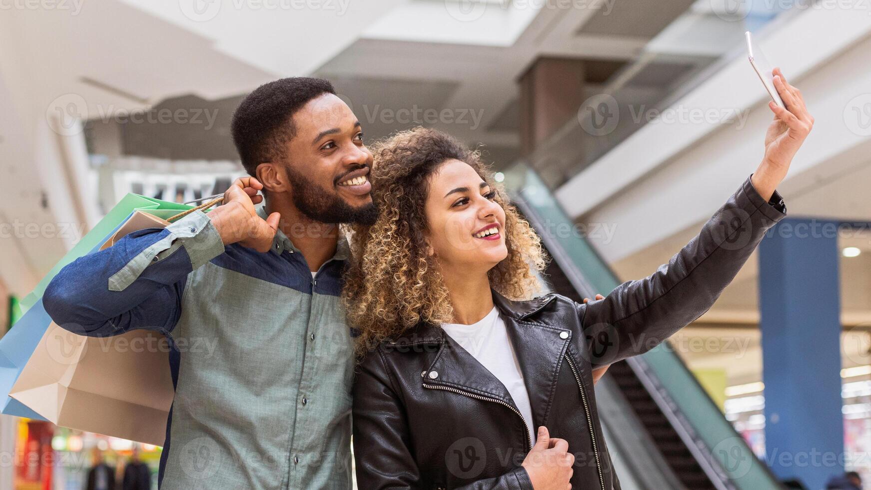 African American man and woman are standing in a bustling shopping mall, holding a smartphone at arms length to take a selfie, smiling and posing in front of a backdrop of stores and shoppers. photo
