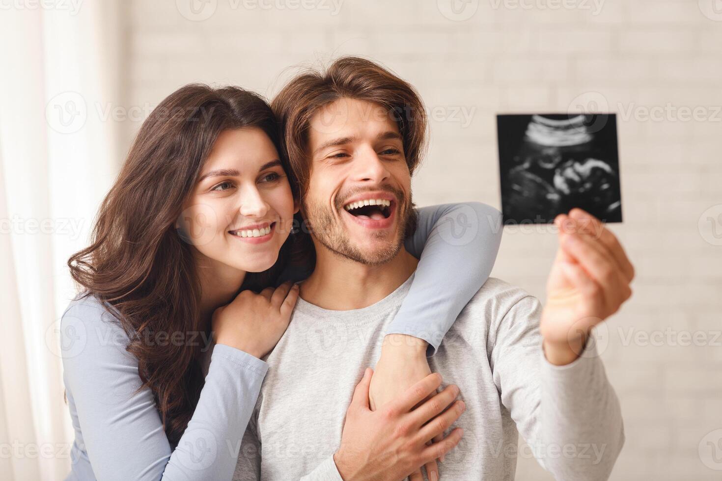 Happy young couple holding and looking at ultrasound scan of their baby, cuddling and bonding at home photo