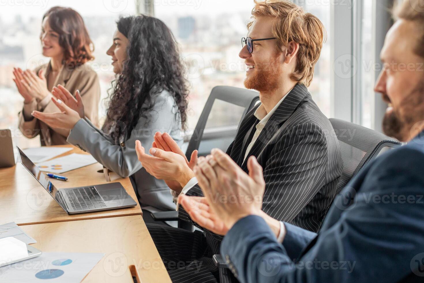 A group of business professionals sit at a table in a modern office, clapping their hands in appreciation as they listen to a presentation. They are dressed in formal attire photo