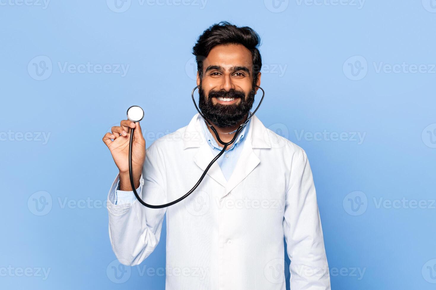 A smiling Indian doctor with a beard holds up a stethoscope against a light blue background. He is wearing a white lab coat and appears confident. He is likely working in the medical field photo