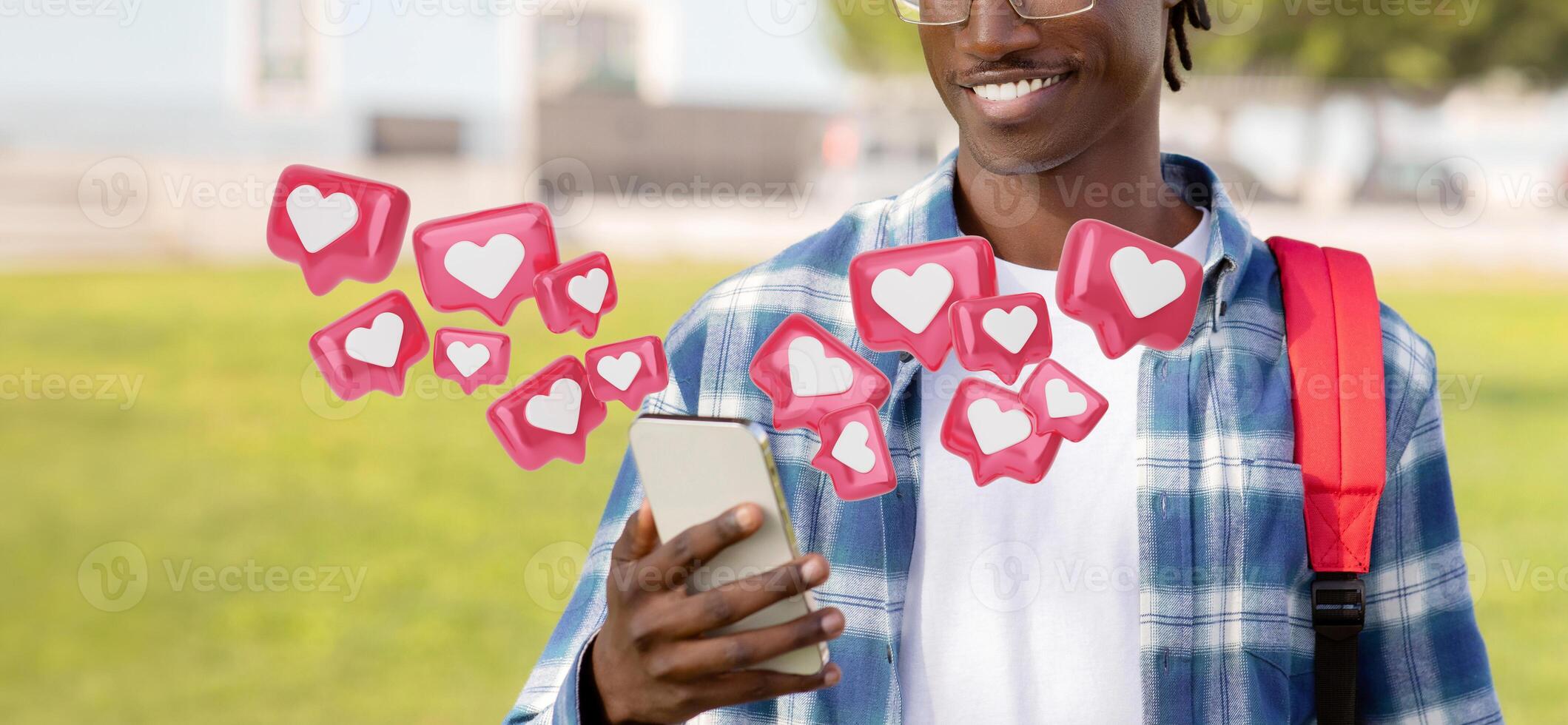 Cropped of young black man wearing a plaid shirt and a backpack is using his smartphone outdoors. He is smiling and looking at his phone screen. Digital hearts are floating around him photo