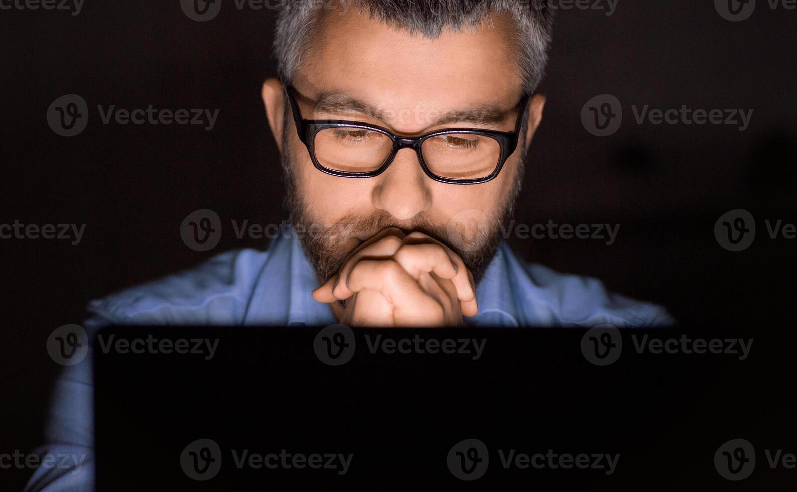 A man with glasses is intently concentrating while working on his laptop in a dimly lit room. His hands are clasped together under his chin as he focuses on the screen. photo