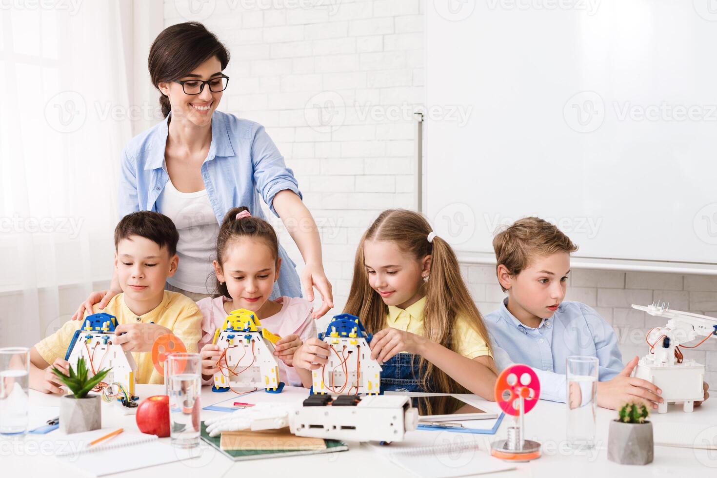 A group of elementary school students are working on a robotics project in a classroom. A teacher is assisting them with their work. The students are learning about how to build and program robots. photo