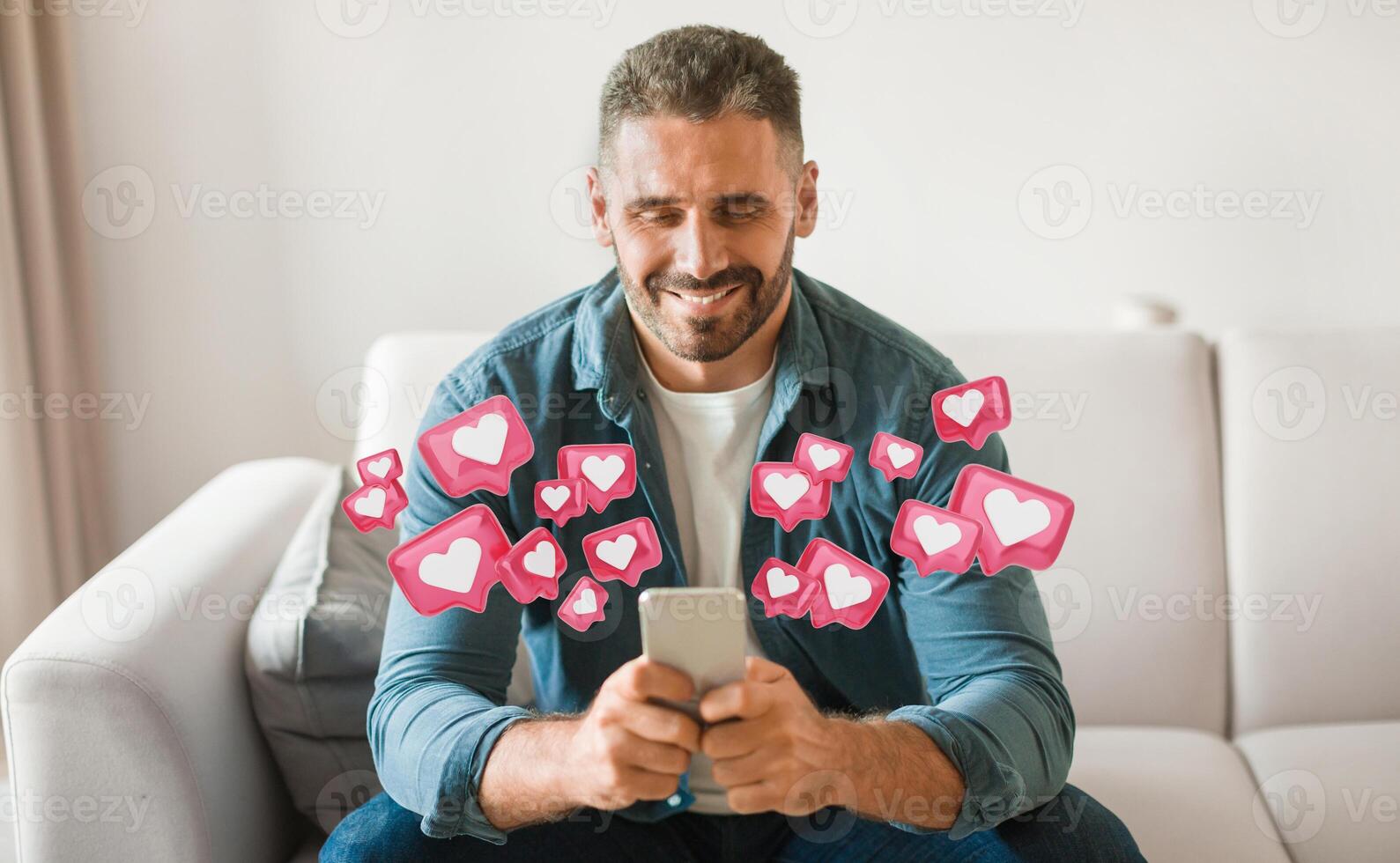 A man is sitting on a white couch, holding a smartphone in his hands, checking his social media. He is smiling as he looks down at his phone. Surrounding him are many pink like icons. photo