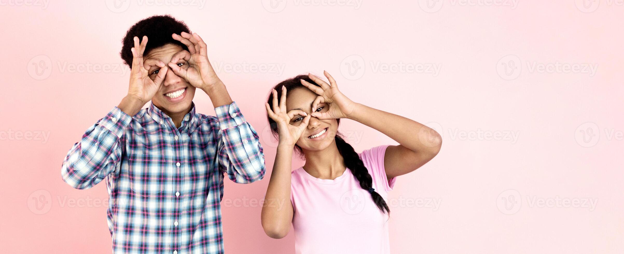 African American guy and girl standing together, both raising their hands up in front of a vibrant pink background. They appear to be in a state of unity or celebration, copy space photo
