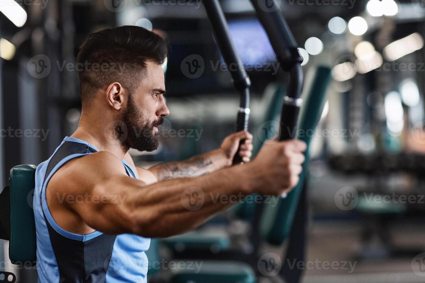 A man with a beard is working out on a chest press machine at the gym. He is focused on his workout and his arms are extended out in front of him. photo