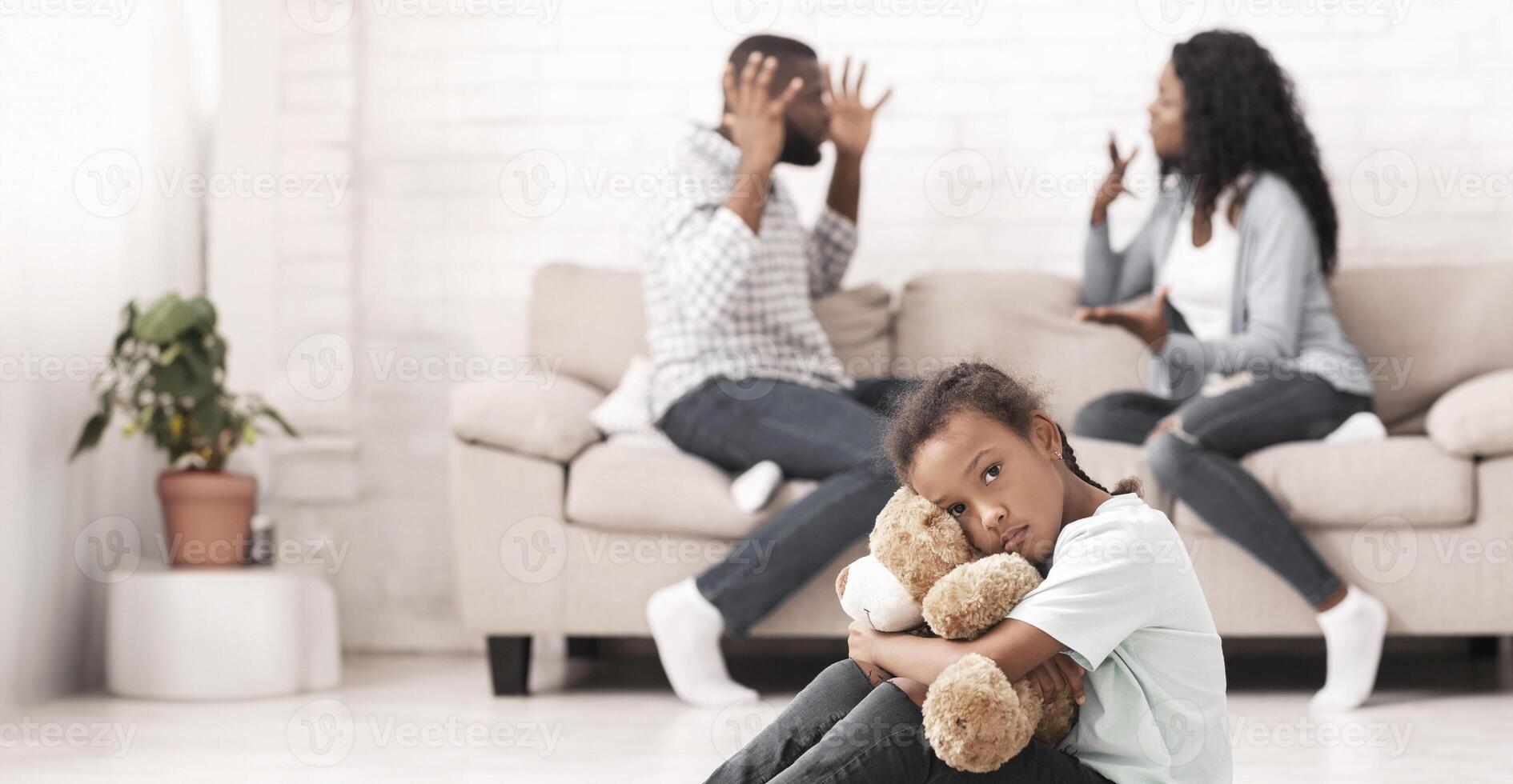 Family problems. Little black girl suffering from parents quarrels, sitting on floor cuddling her teddy bear, feeling lonely and depressed photo