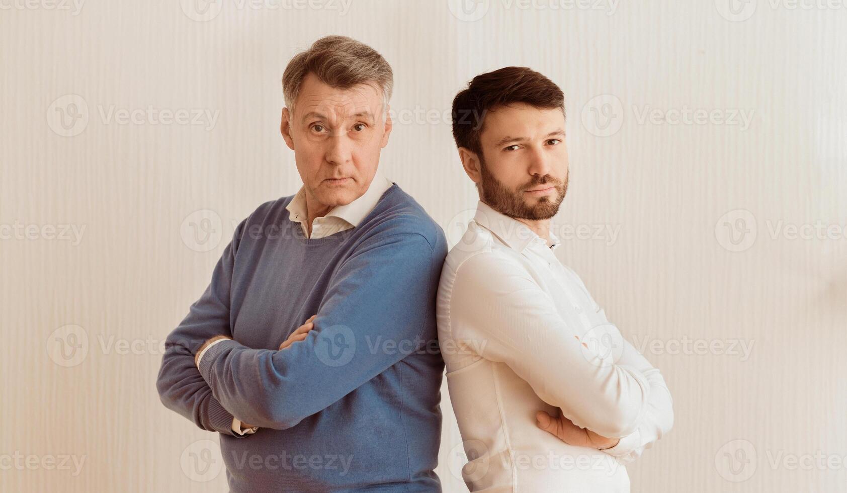 Relationship Problems. Serious Adult Son And Senior Father Standing Back-To-Back Crossing Hands Against White Wall Indoor photo