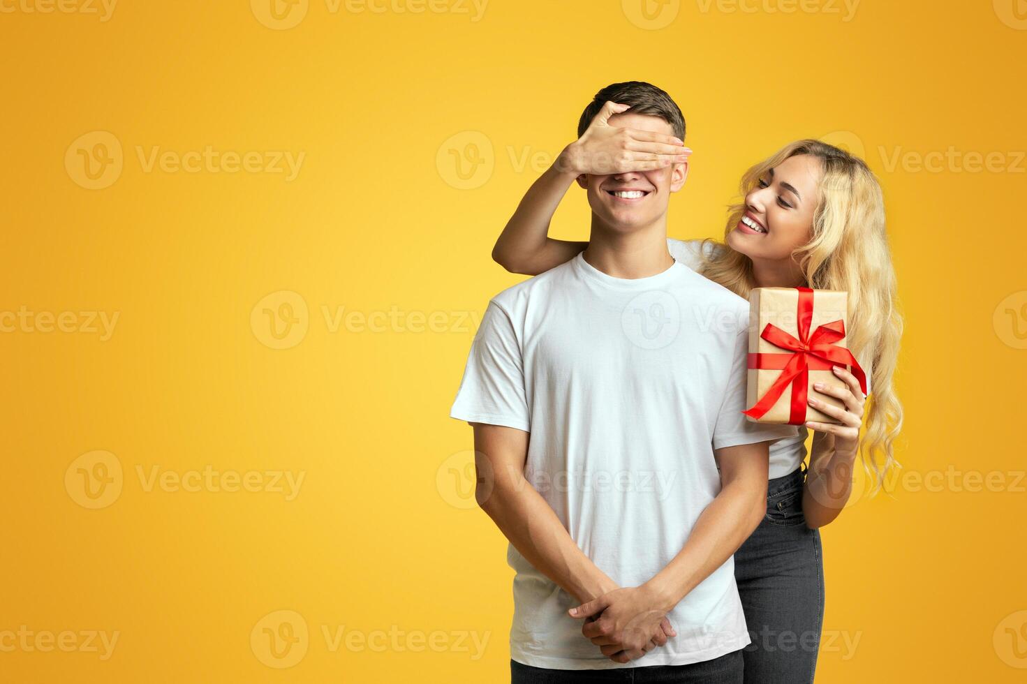 Surprise. Loving girl prepared romantic present to her boyfriend and covering his eyes while standing against yellow background in studio, copy space photo