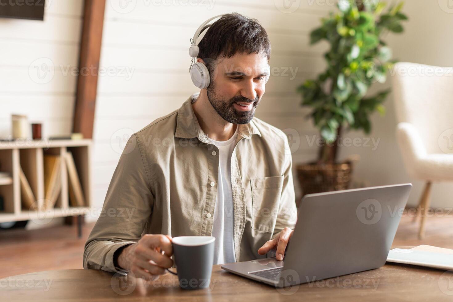 A young man is seated at a wooden table, engaging in remote work on his laptop. He wears headphones and holds a mug, suggesting a comfortable and casual work environment photo