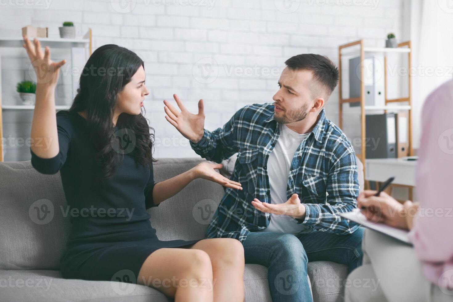 A young couple sits on a couch in a therapists office, engaged in a heated argument. The woman is gesturing with her hands, while the man is seated with a serious expression on his face. photo