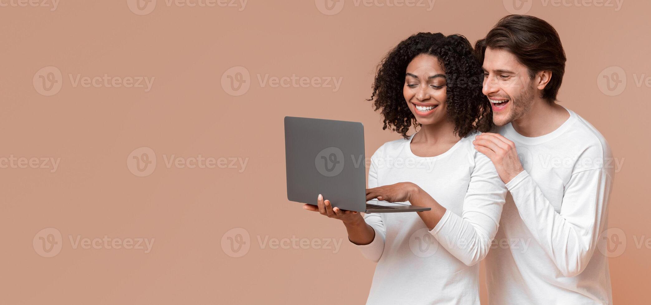Online sales concept. Happy interracial spouses holding laptop and looking at computer screen with excitement, enjoying internet shopping photo