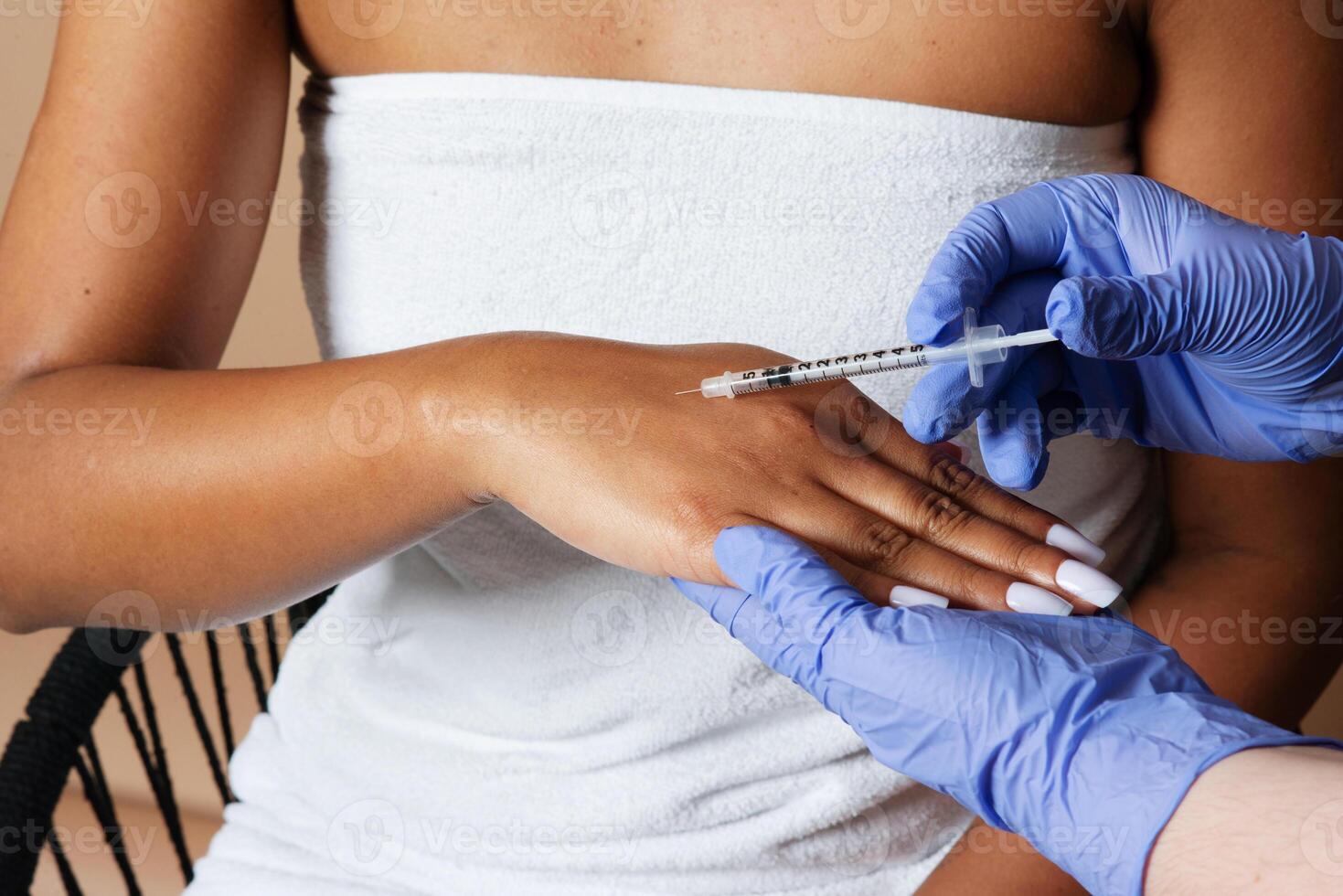 A medical professional is administering a syringe injection into the hand of a woman. The woman appears to be calmly receiving the medical procedure in a clinical setting, cropped photo