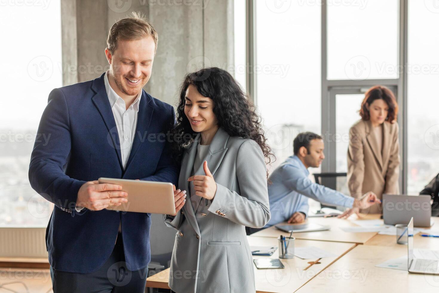 Two business professionals are working together in a modern office setting. They are standing near a large wooden table with other colleagues in the background. The man is holding a tablet photo