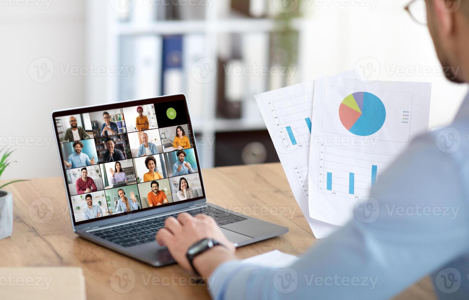 A businessman sits at a desk in his office, using a laptop to participate in a virtual meeting with colleagues. The screen shows a grid of calls with many diverse people photo
