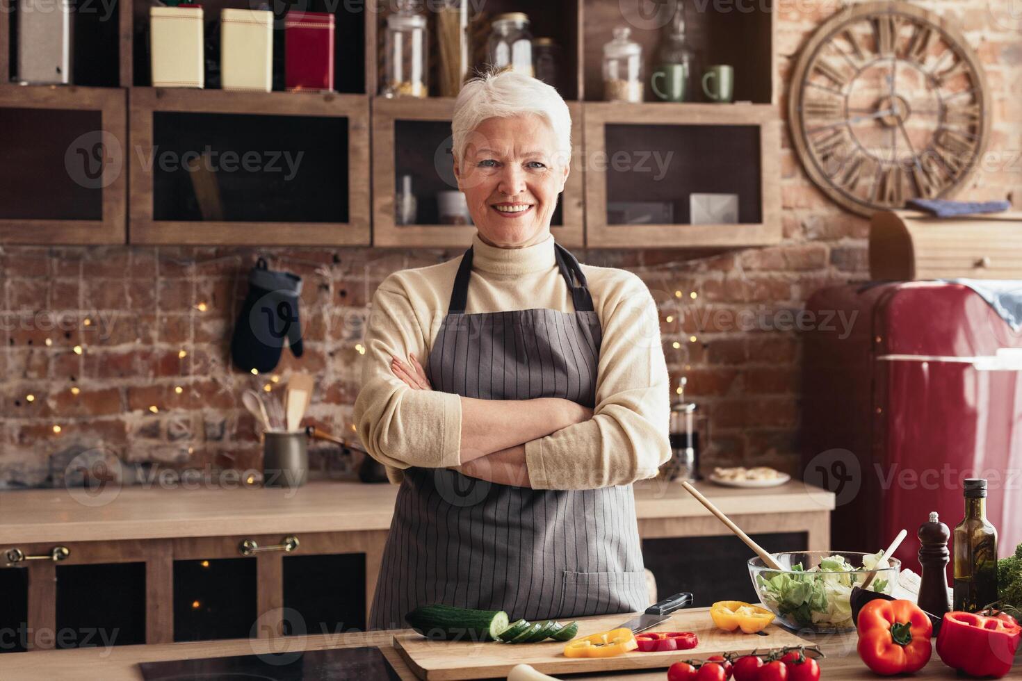 A mature woman with short gray hair smiles while standing in her kitchen with her arms crossed. She is wearing an apron and there are various cooking ingredients and utensils on the countertop. photo