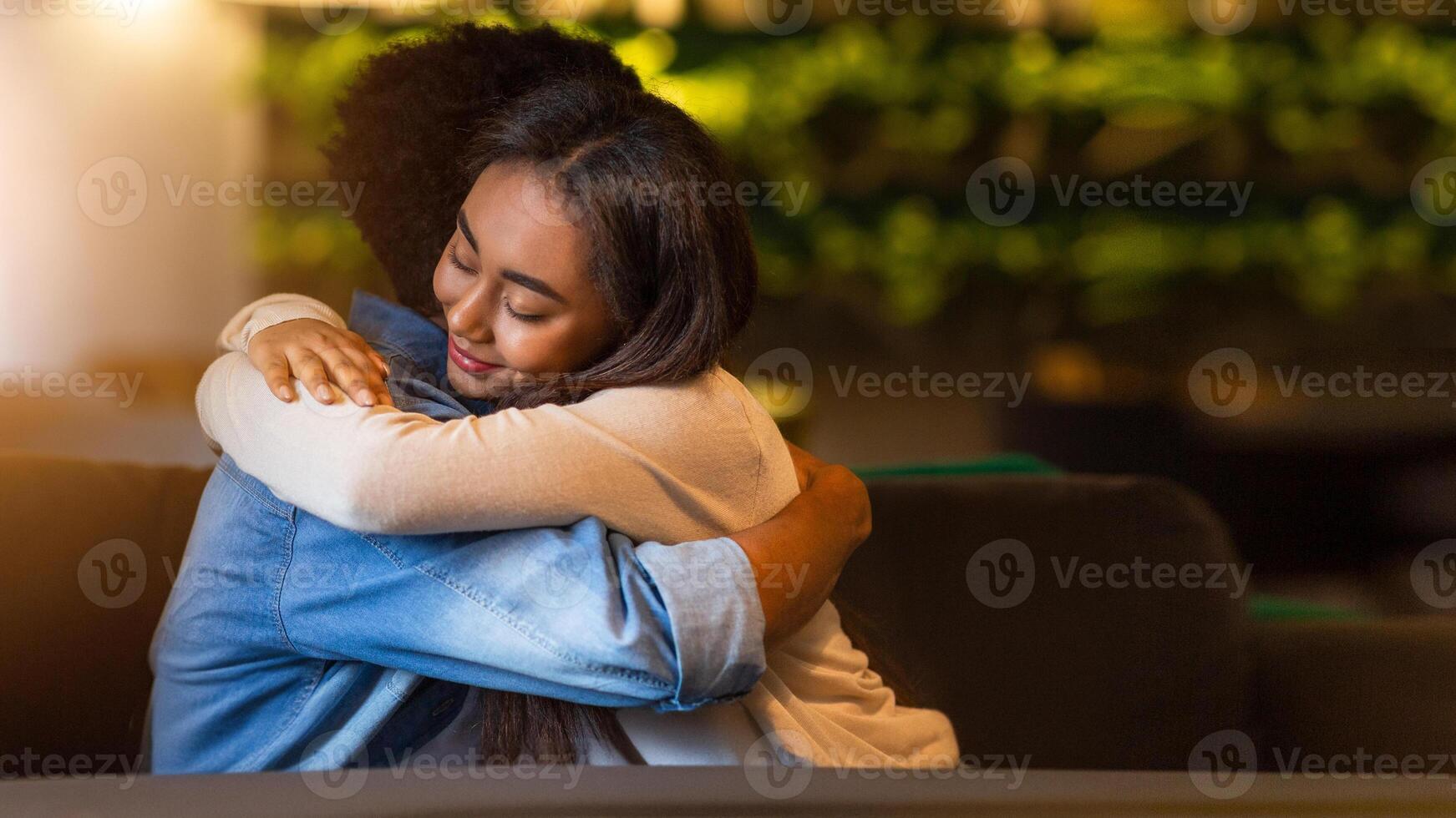 Two black friends share a heartfelt embrace inside a cozy cafe with soft, dim lighting. The background features lush, green decor which adds to the warm atmosphere of the evening setting. photo