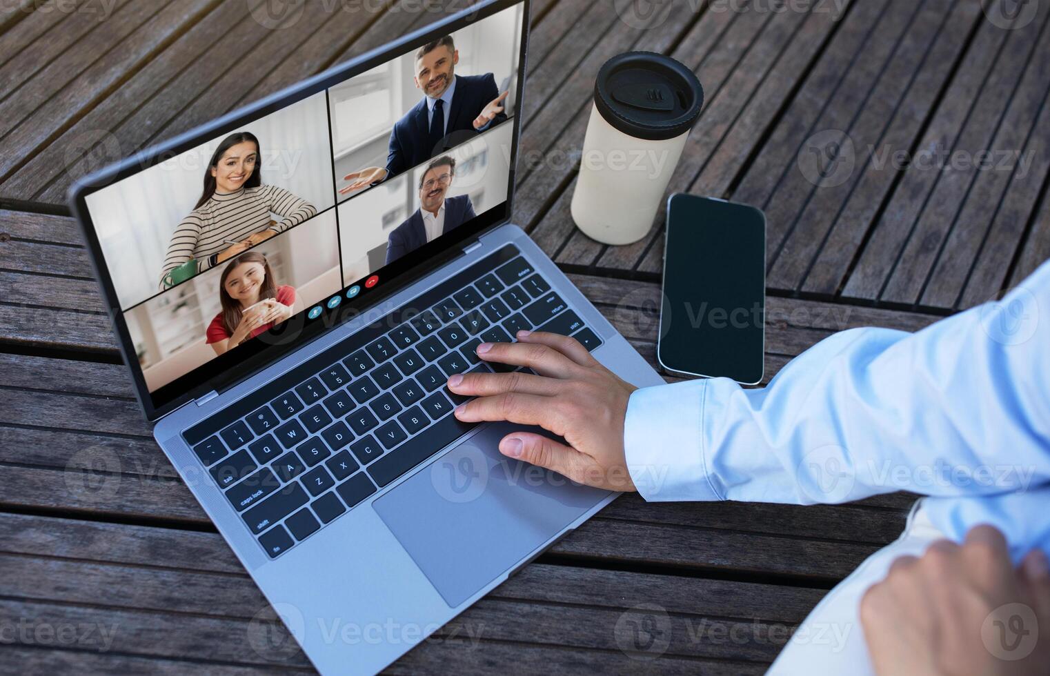 A person is sitting outdoors at a table and using a laptop to participate in a conference call. The laptop screen shows multiple people on a call. photo