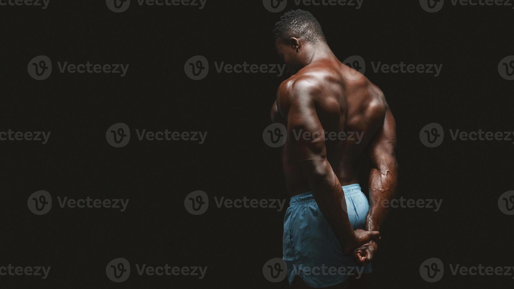 A muscular man stands with his back to the camera, showing off his physique against a dark background. photo