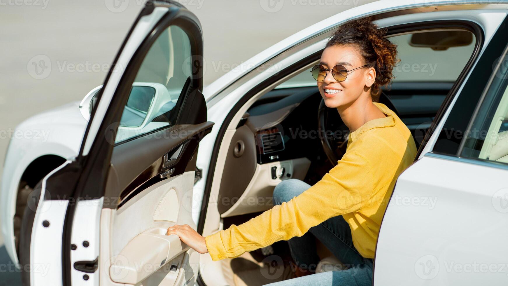 A young black woman wearing a yellow shirt and sunglasses gets out of a white car on a sunny day. Her hair is pulled back in a messy bun. The car door is open and she is leaning out of it. photo