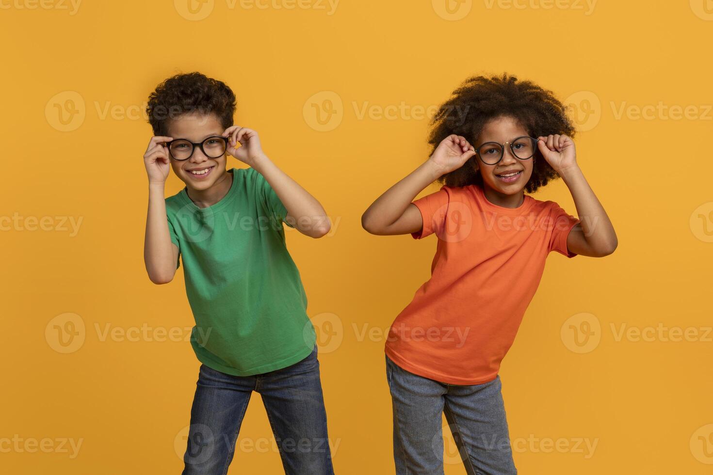 Two African American children, one wearing a green shirt and the other an orange shirt, pose for a portrait against a yellow background. They both wear glasses and look at the camera with smiles. photo