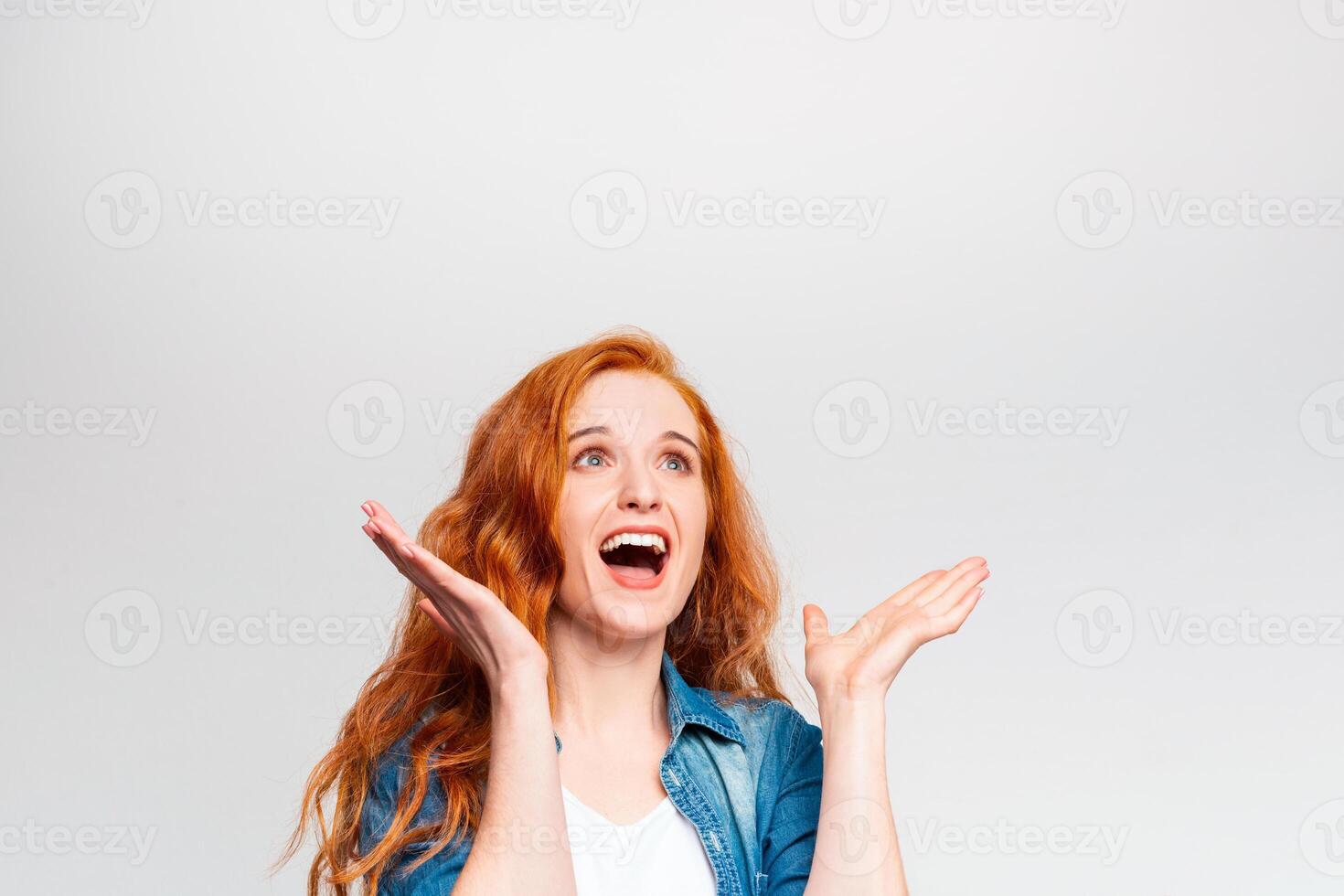 A woman with bright red hair and a denim jacket is looking upwards with an open-mouthed smile, her arms raised in a cheerful and surprised gesture, copy space photo