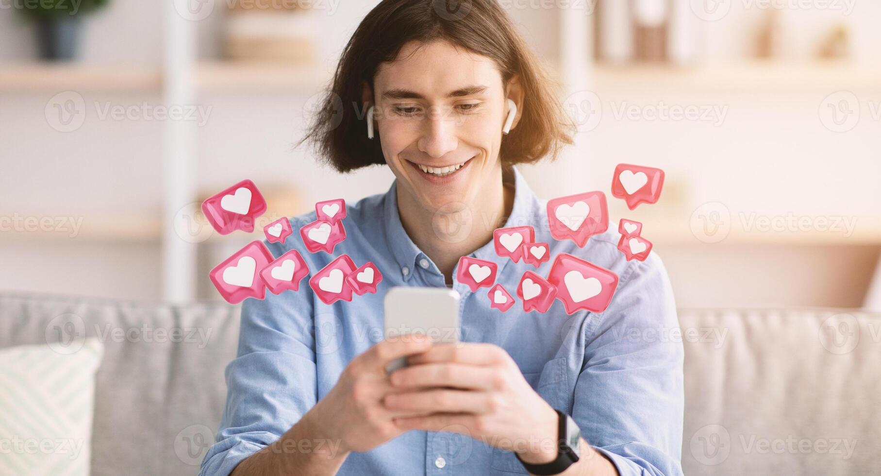 Young man sits on couch, smiling, as he uses his smartphone. He is surrounded by animated pink hearts with white interiors. This image depicts the joy of receiving positive feedback on social media. photo