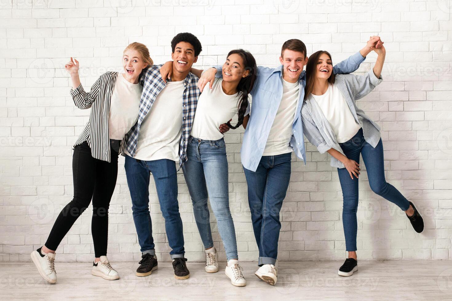 A group of five multiethnic teenagers are standing in a row against a white brick wall, smiling and laughing. They are dressed in casual clothing photo