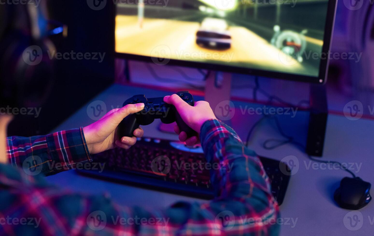 This image shows a gamer playing a racing game at their computer desk. The gamer is wearing a plaid shirt and using a black game controller. The computer monitor is displaying racing game photo
