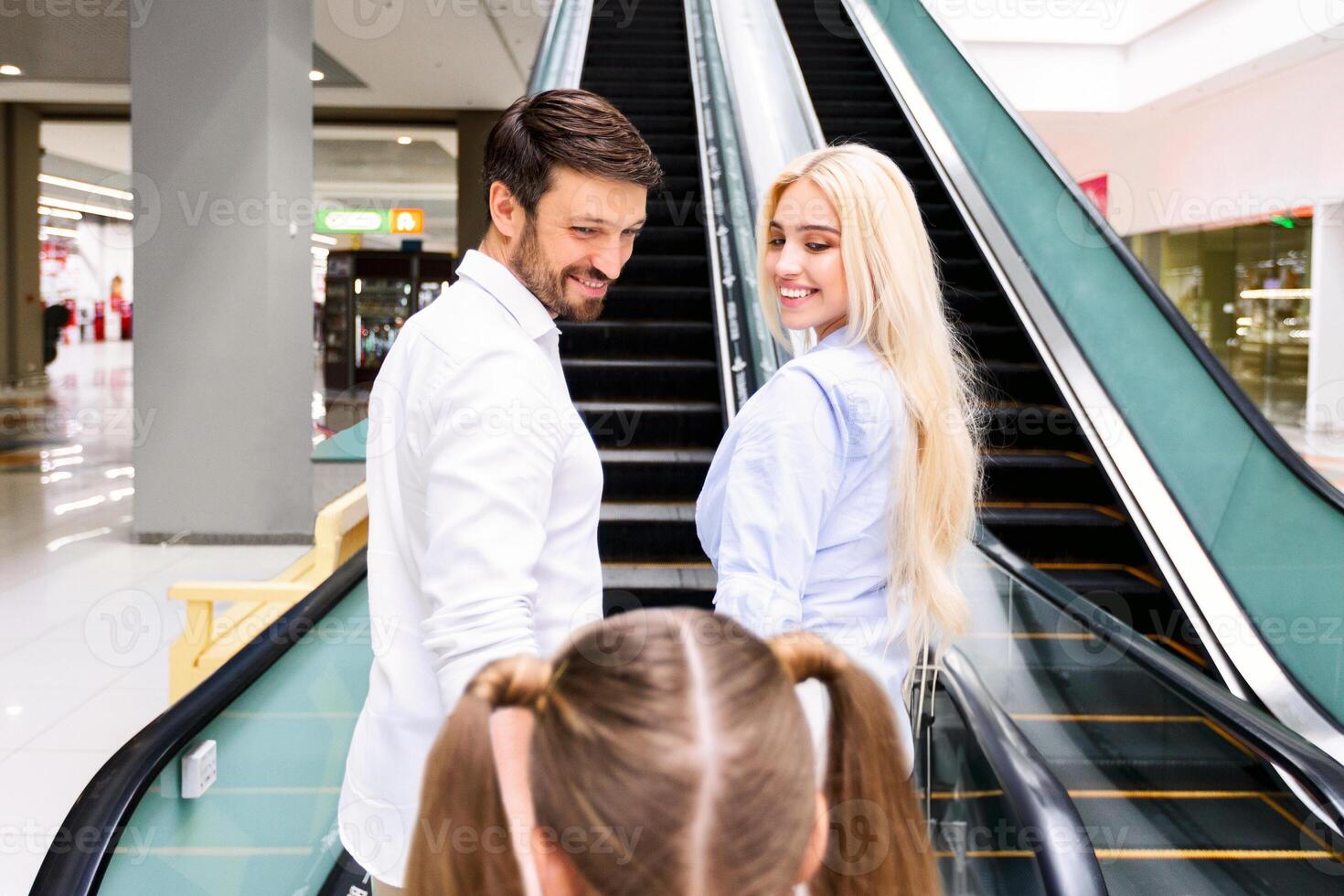 A family of three rides an escalator up in a large shopping mall. The parents are in the foreground, while their young daughter stands in front of them, looking at the camera. photo