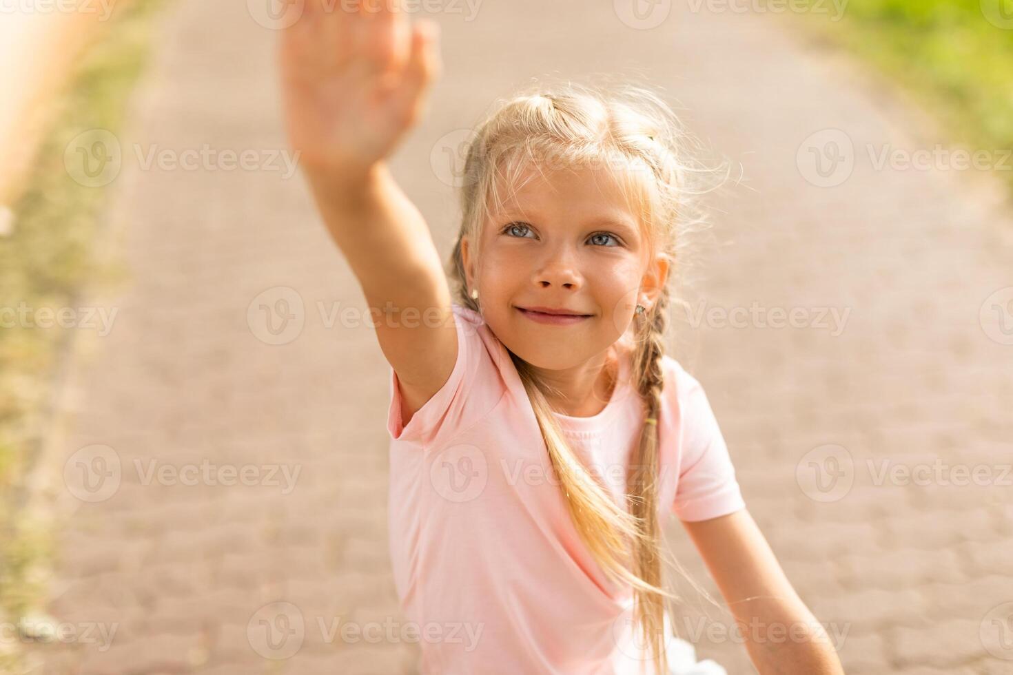A young girl with blonde hair and blue eyes is shown reaching out with her right hand. She is wearing a light pink t-shirt and is looking directly at the camera with a warm smile, closeup photo