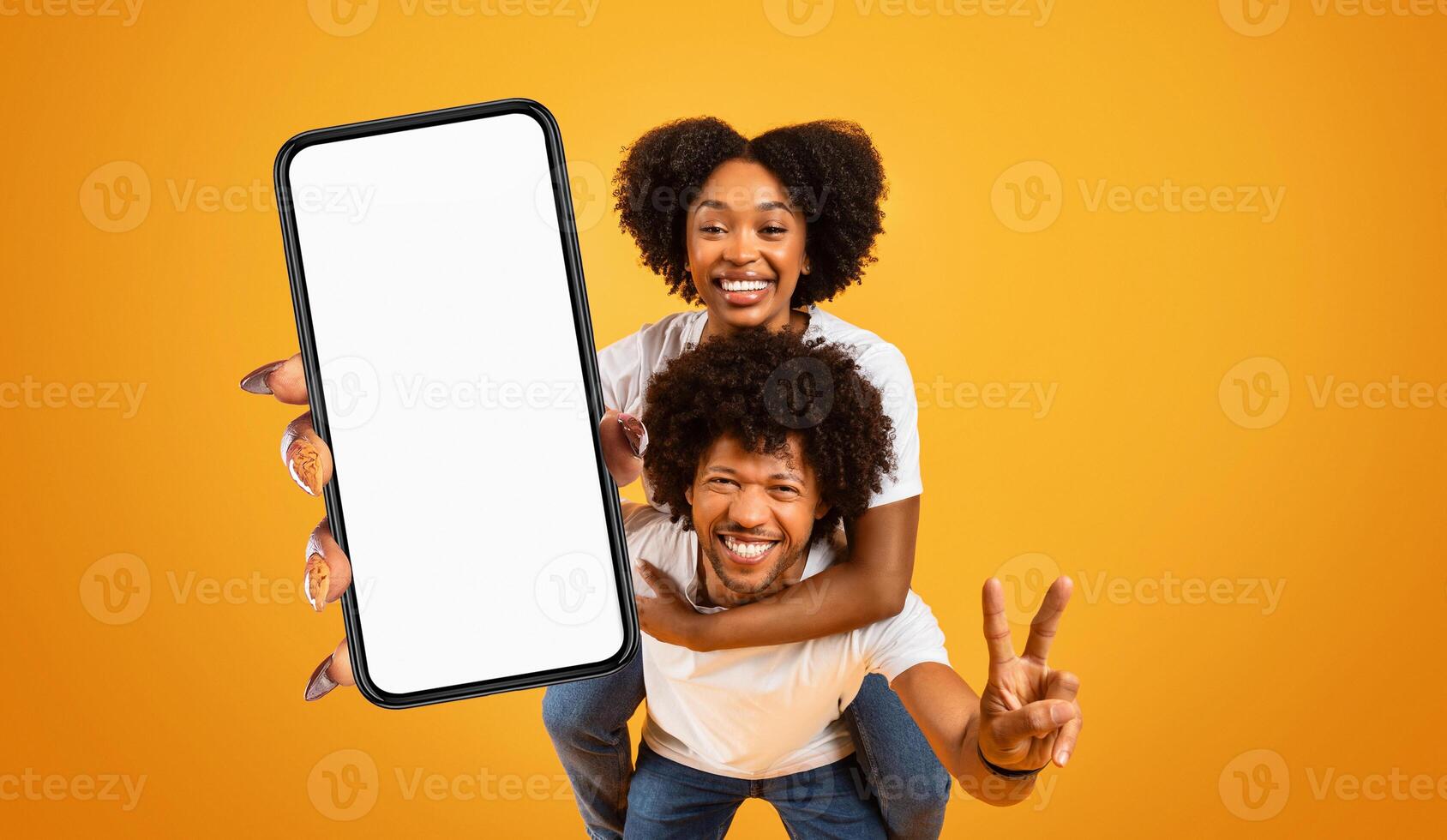 A happy black couple is holding a smartphone with a blank screen in front of a bright yellow background. The woman is giving a peace sign with her right hand and is smiling brightly. photo