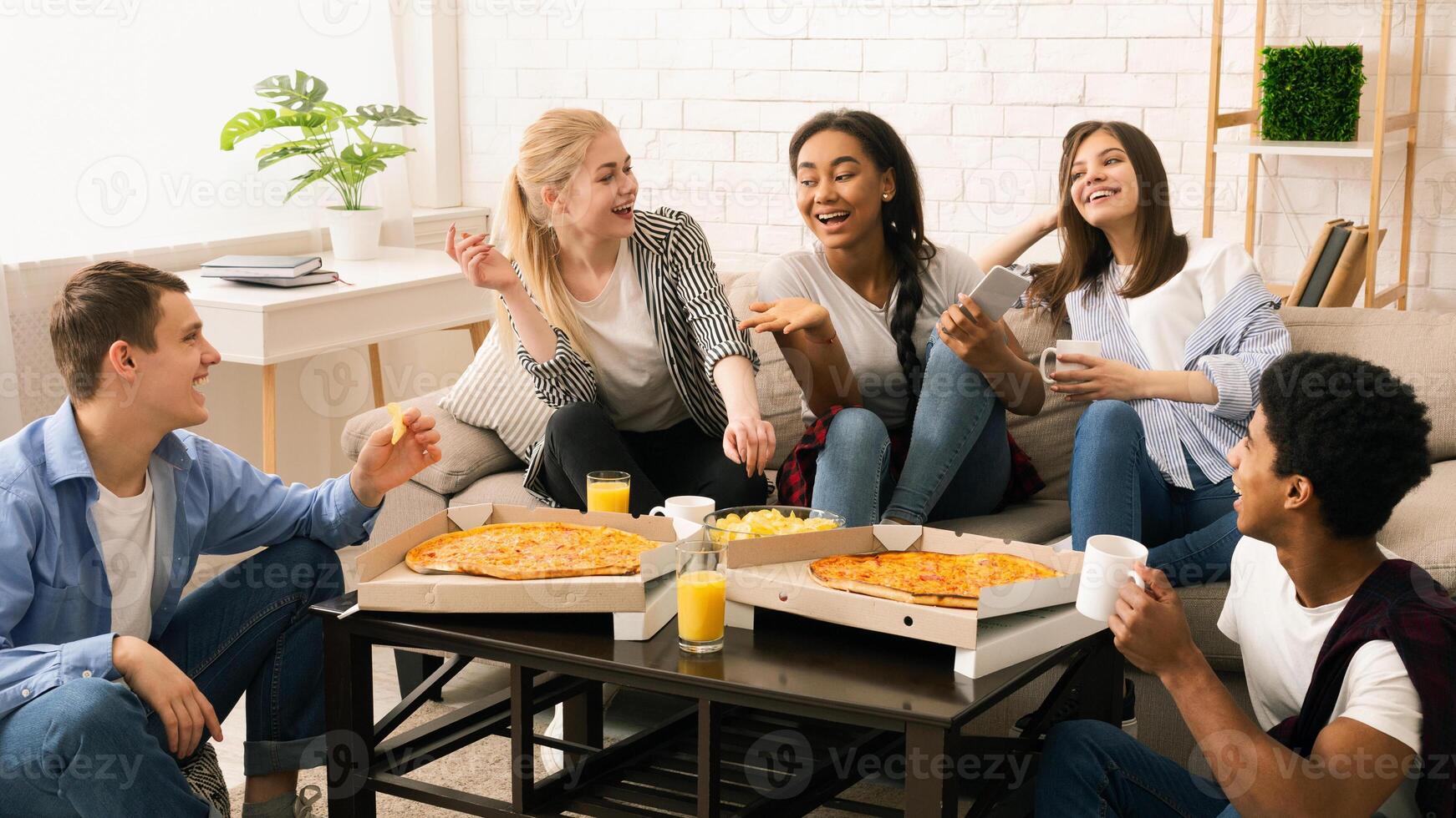 A diverse group of multiethnic friends are gathered around a wooden table. They are enjoying slices of hot pizza, engaged in casual conversation and laughter. photo