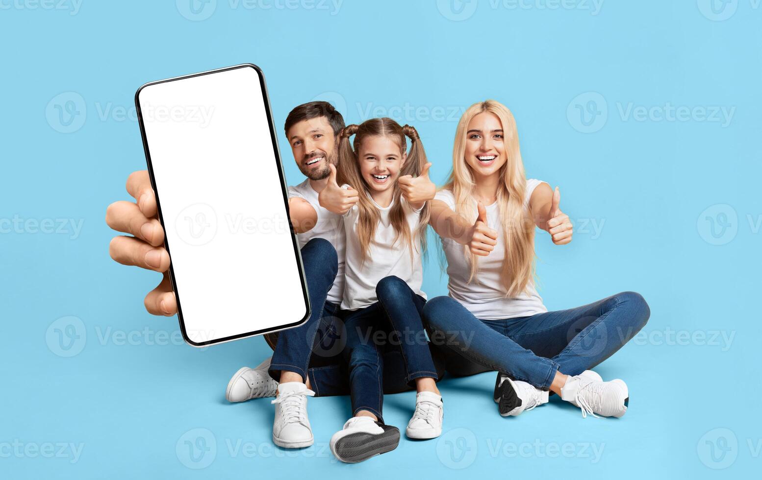 A happy family of three is sitting on the floor against a light blue background. They are holding up a large smartphone with a blank screen and giving thumbs up. photo
